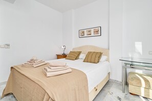 A neatly made bed with a beige bedspread, two pillows, and folded towels. There is a bedside table with a lamp, a framed picture on the wall, and a glass table beside the bed.