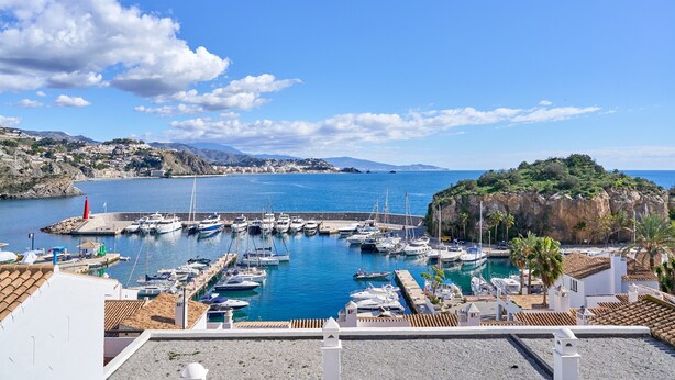 Un port de plaisance avec des bateaux amarrés, un ciel bleu azur et une chaîne de montagnes en arrière-plan.