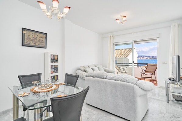 A modern living room with a glass dining table, grey sofa, and a balcony with a view.