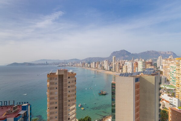 Eine Küstenstadtlandschaft mit Hochhäusern, freiem Blick auf das Meer und Bergen im Hintergrund.