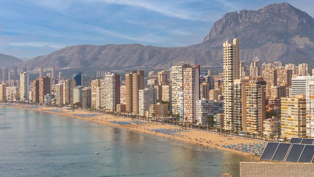 Paesaggio costiero con edifici alti, una spiaggia e montagne sullo sfondo.