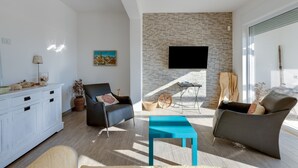 A modern living room with a stone wall, a flat-screen TV, and a dining area with a table and chairs. - Villa "Domus Bianca" with Sea View, Garden & Wi-Fi (San Giovanni di Sinis)