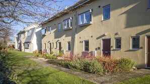 A row of townhouses with individual front doors, a well-maintained lawn, and a clear sky. - Clearwater 31, The Lakehouse - Waterside Holiday Cottage, Spa Access, Sleeps 8 (Somerford Keynes)