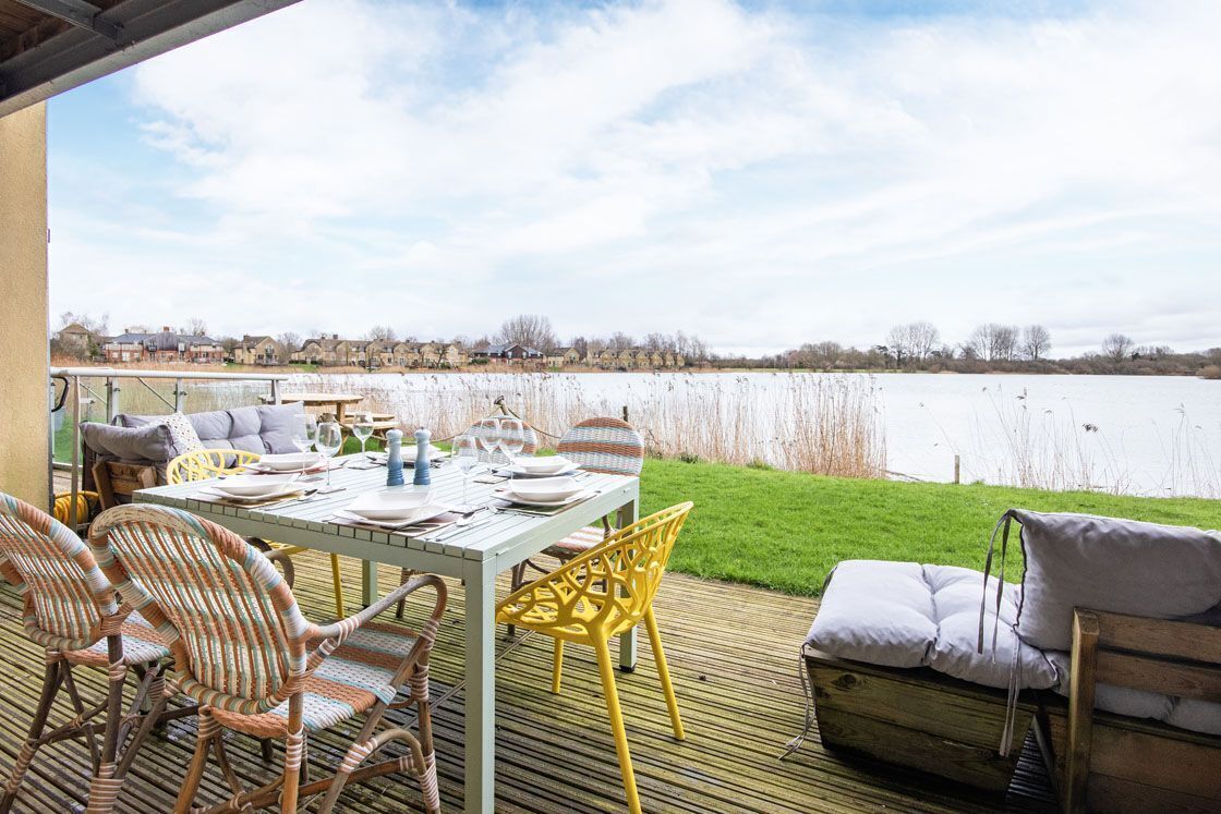 Un área de comedor al aire libre con una mesa preparada para cuatro, sillas de mimbre y una terraza de madera con vista a un cuerpo de agua.