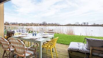 Un área de comedor al aire libre con una mesa preparada para cuatro, sillas de mimbre y una terraza de madera con vista a un cuerpo de agua.