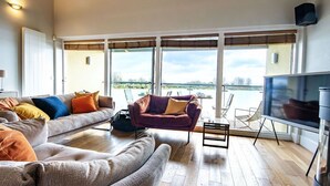 A spacious living room with a large sofa, a smaller armchair, and a television. - Clearwater 31, The Lakehouse - Waterside Holiday Cottage, Spa Access, Sleeps 8 (Somerford Keynes)