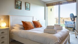 A neatly made bed with white and orange bedding, a bedside lamp, and a view of the outdoors through a large window. - Clearwater 31, The Lakehouse - Waterside Holiday Cottage, Spa Access, Sleeps 8 (Somerford Keynes)