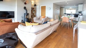 A modern living room with a sofa, a TV, a fireplace, and a dining area with chairs and a table. - Clearwater 31, The Lakehouse - Waterside Holiday Cottage, Spa Access, Sleeps 8 (Somerford Keynes)