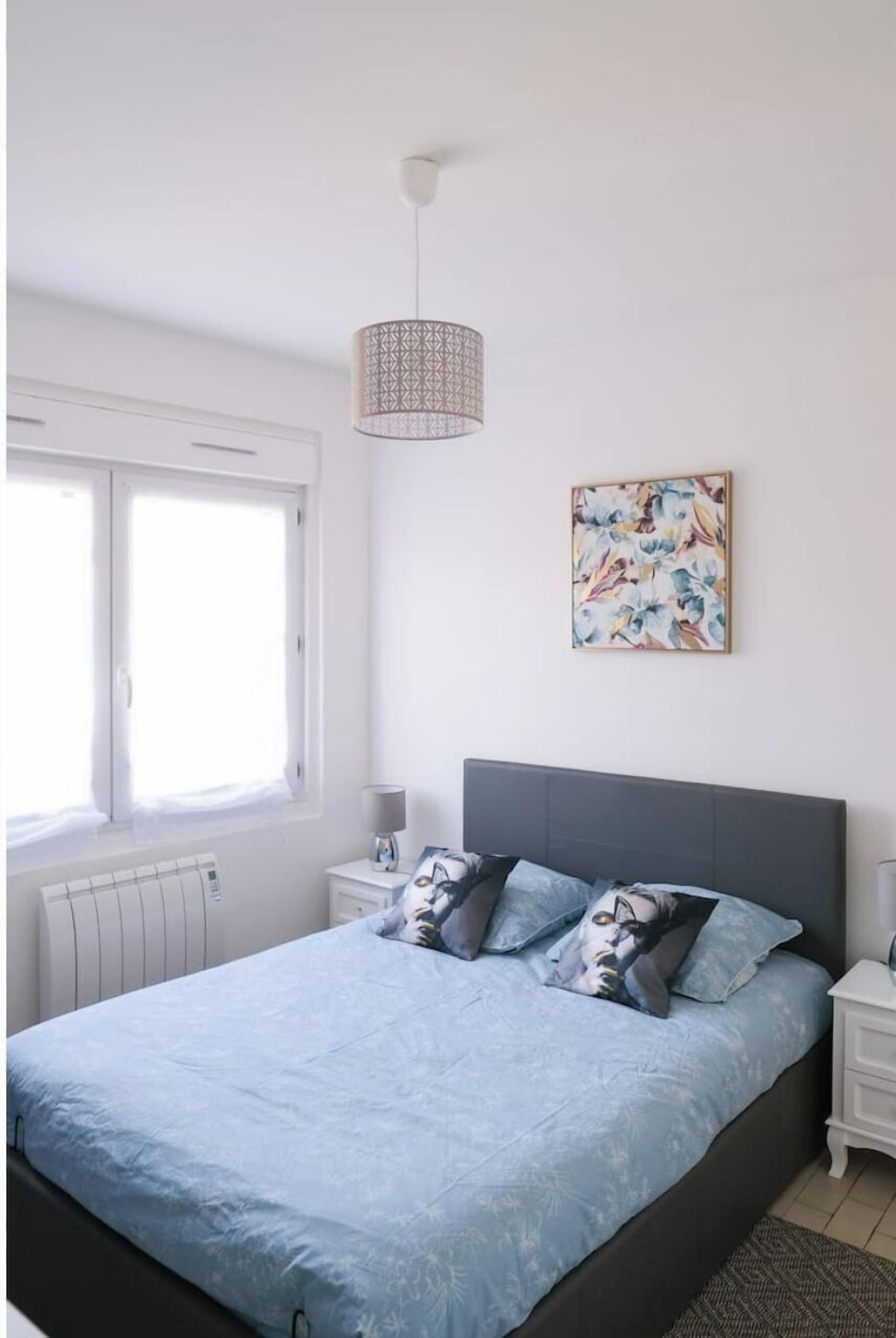 A bedroom with a bed, a bedside table, a lamp, a window, and a wall-mounted artwork.
