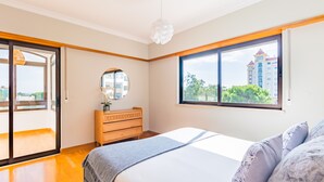Ein Schlafzimmer mit einem Bett, einer Kommode, einem Spiegel und einem Fenster mit Blick auf Gebäude und Bäume.