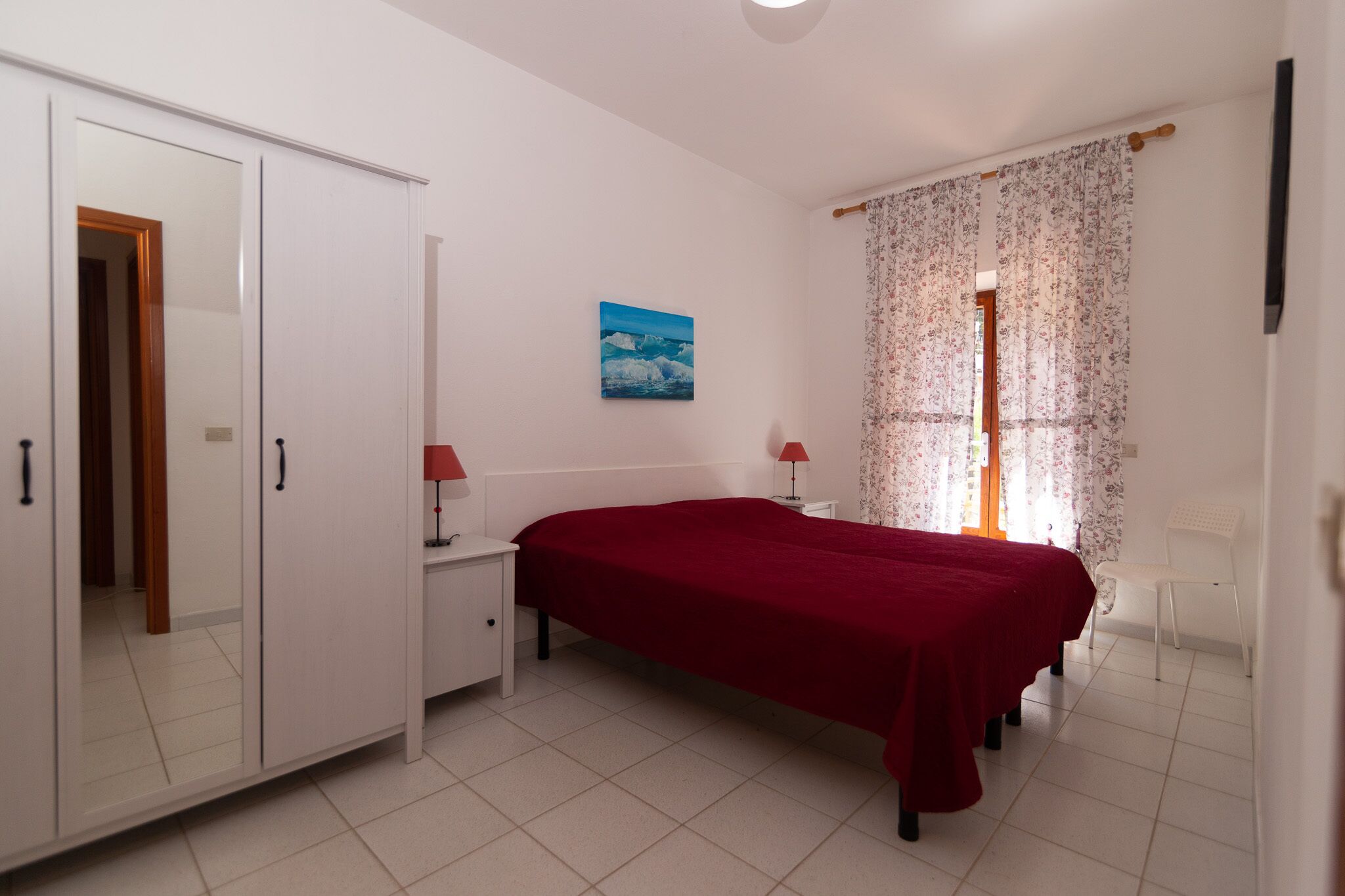 A bedroom with a bed, a wardrobe, a nightstand, a lamp, a chair, and a window with curtains.