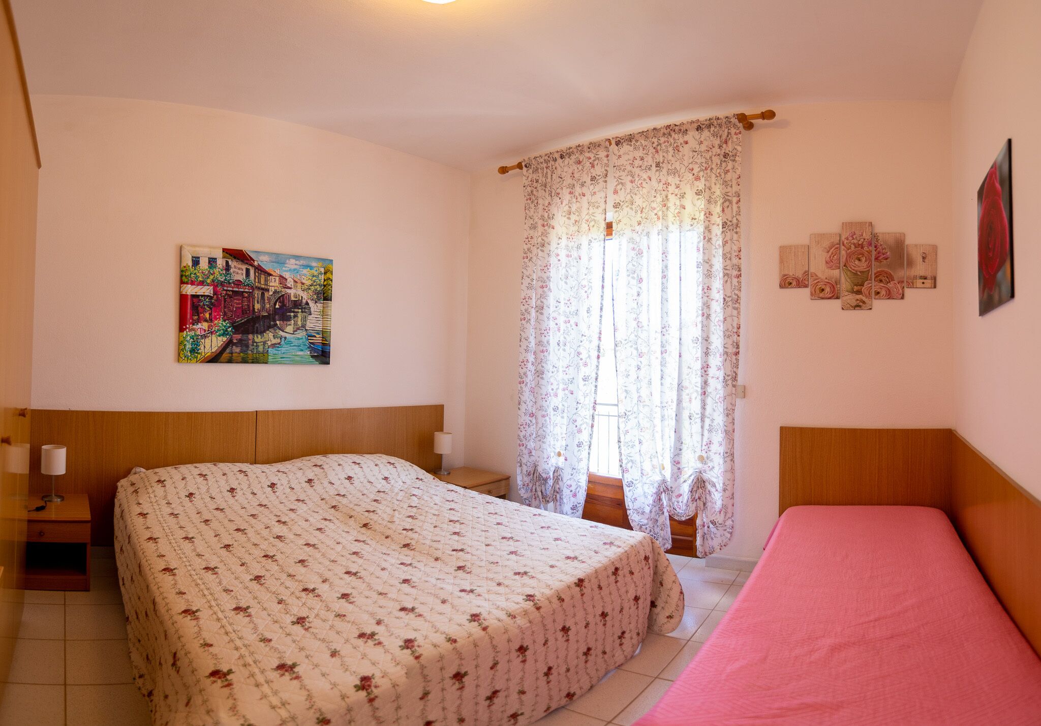 Un dormitorio con una cama grande, una cama más pequeña, una mesita de noche, una lámpara, una ventana con cortinas florales y un cuadro en la pared.