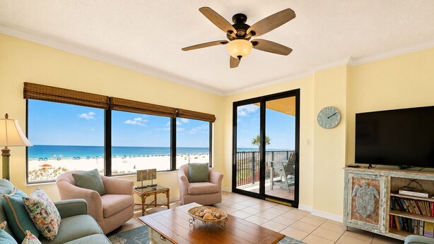 A living room with a view of the beach, a ceiling fan, a flat-screen TV, and a bookshelf. - Private Fishing Pier! (Orange Beach)