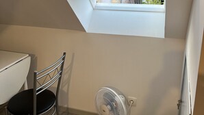 Une petite pièce avec un plafond en pente, une fenêtre, une table, une chaise et un ventilateur sur pied.