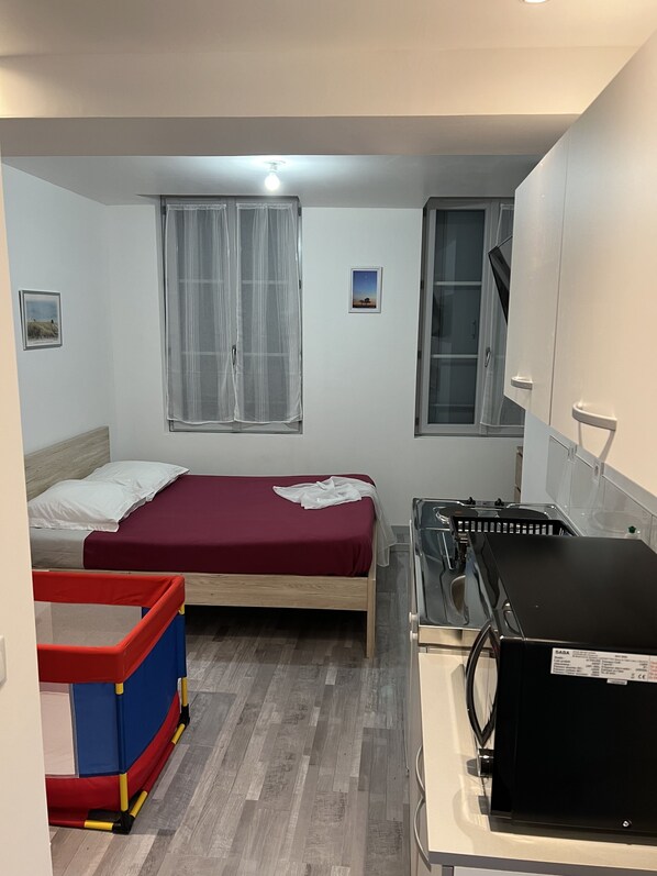 A compact room with two beds, a small kitchenette, a window with sheer curtains, and a child's playpen.