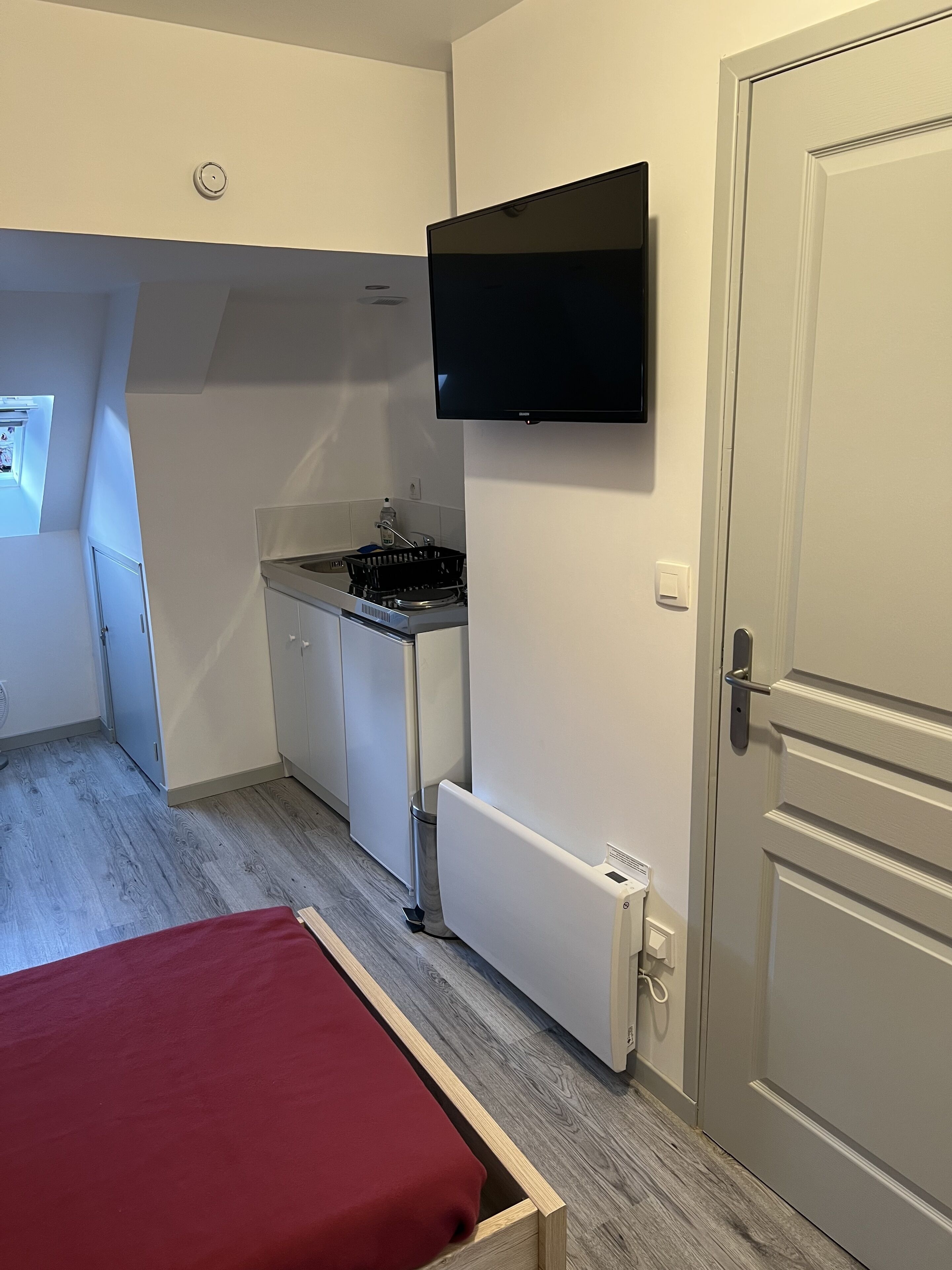 Habitación compacta con kitchenette, cama con cubierta roja y televisor montado en la pared.