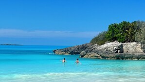 Ein Strand mit klarem türkisblauem Wasser, einem Felsen mit Vegetation und einer entfernten Insel.