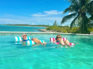 Deux personnes dans une piscine à l’eau cristalline, avec vue sur l’océan et les palmiers.