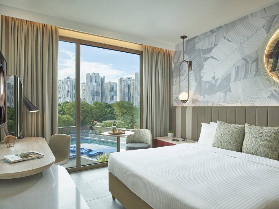 Una habitación de hotel moderna con una cama grande, un escritorio con una computadora portátil, una silla, una mesita con una bebida y vistas a la silueta urbana a través de amplios ventanales.