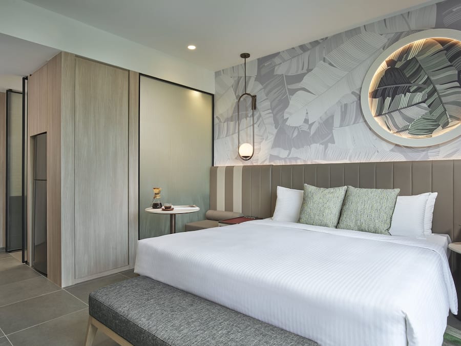 A modern hotel room with a large bed, a round wall decoration, a small table with a lamp, and a bench.