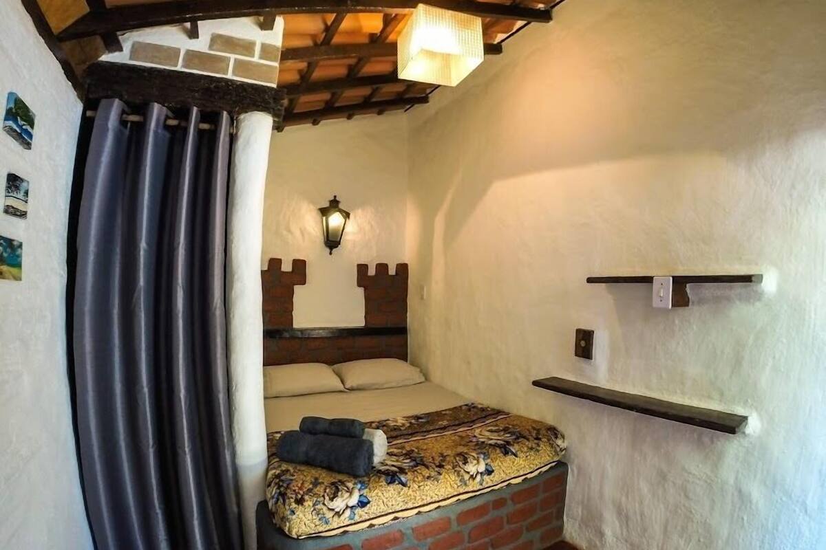 Quarto com cabeceira de tijolos, cama com colcha estampada, luminária pendente e uma prateleira pequena.