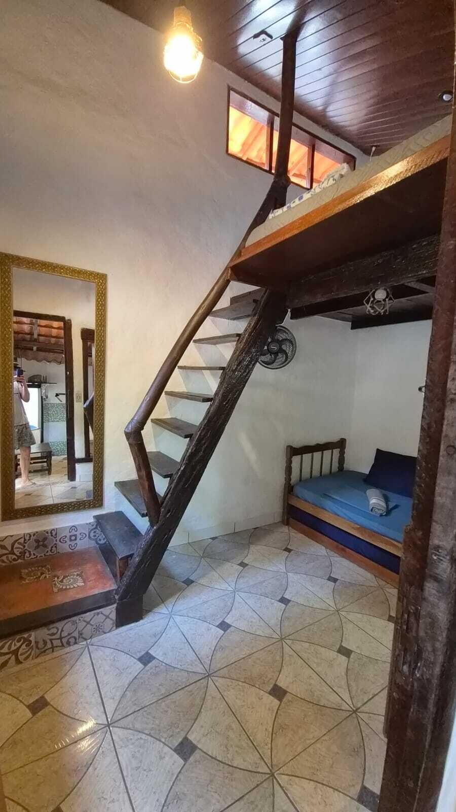Una habitación con techo inclinado, escalera de madera, cama con colchón azul y suelo de baldosas.