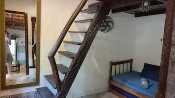 Una habitación con techo inclinado, escalera de madera, cama con colchón azul y suelo de baldosas.