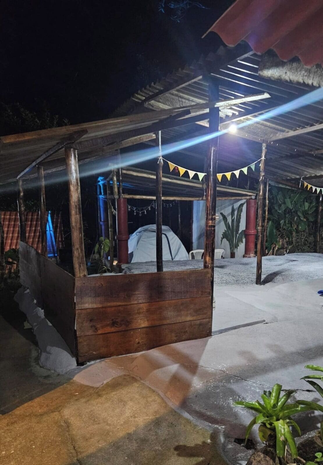 A covered outdoor seating area with a wooden bench, a tent in the background, and a string of lights.