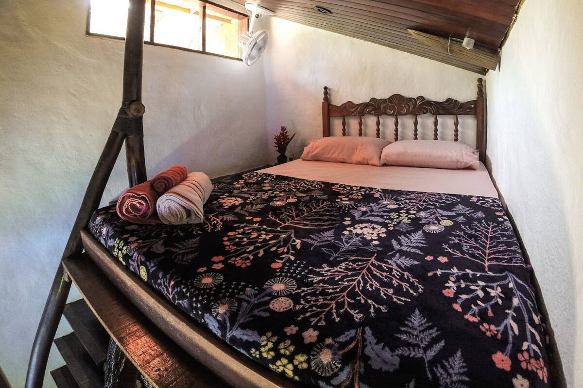 Uma cama de solteiro com colcha florida, cabeceira de madeira e toalhas dobradas em uma cadeira de madeira.