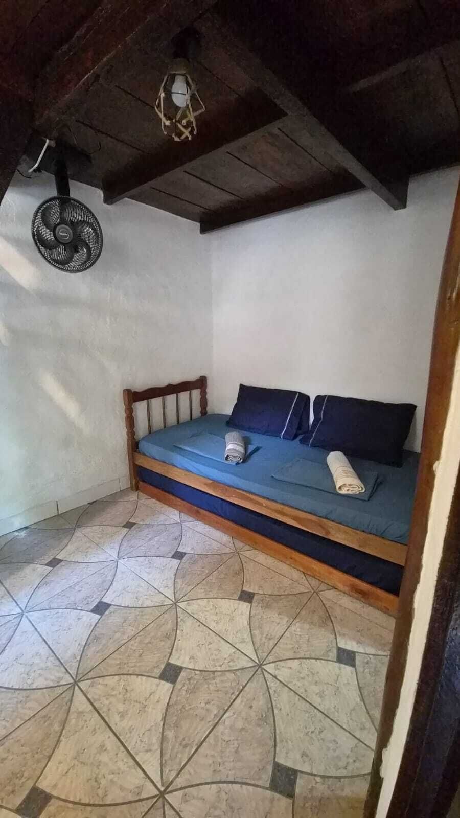 Une petite chambre avec un lit simple, un cadre en bois, un matelas bleu et un ventilateur au plafond.