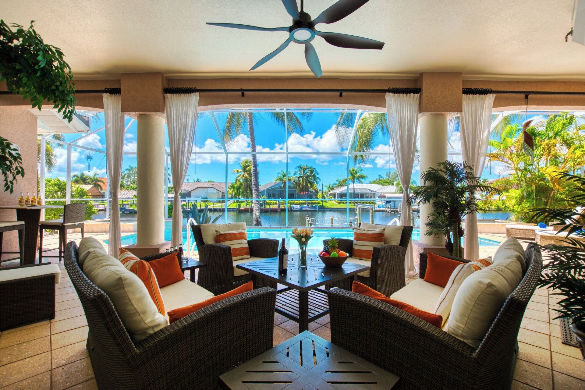 A sunlit outdoor lounge area with wicker furniture, a ceiling fan, and a view of a swimming pool and tropical landscape.