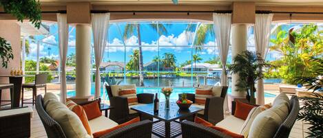A sunlit outdoor lounge area with wicker furniture, a ceiling fan, and a view of a swimming pool and tropical landscape.
