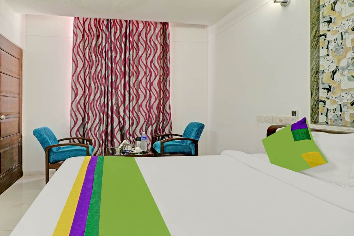 Quarto de hotel com cama, duas poltronas, uma mesa com garrafa de água e cortina estampada.