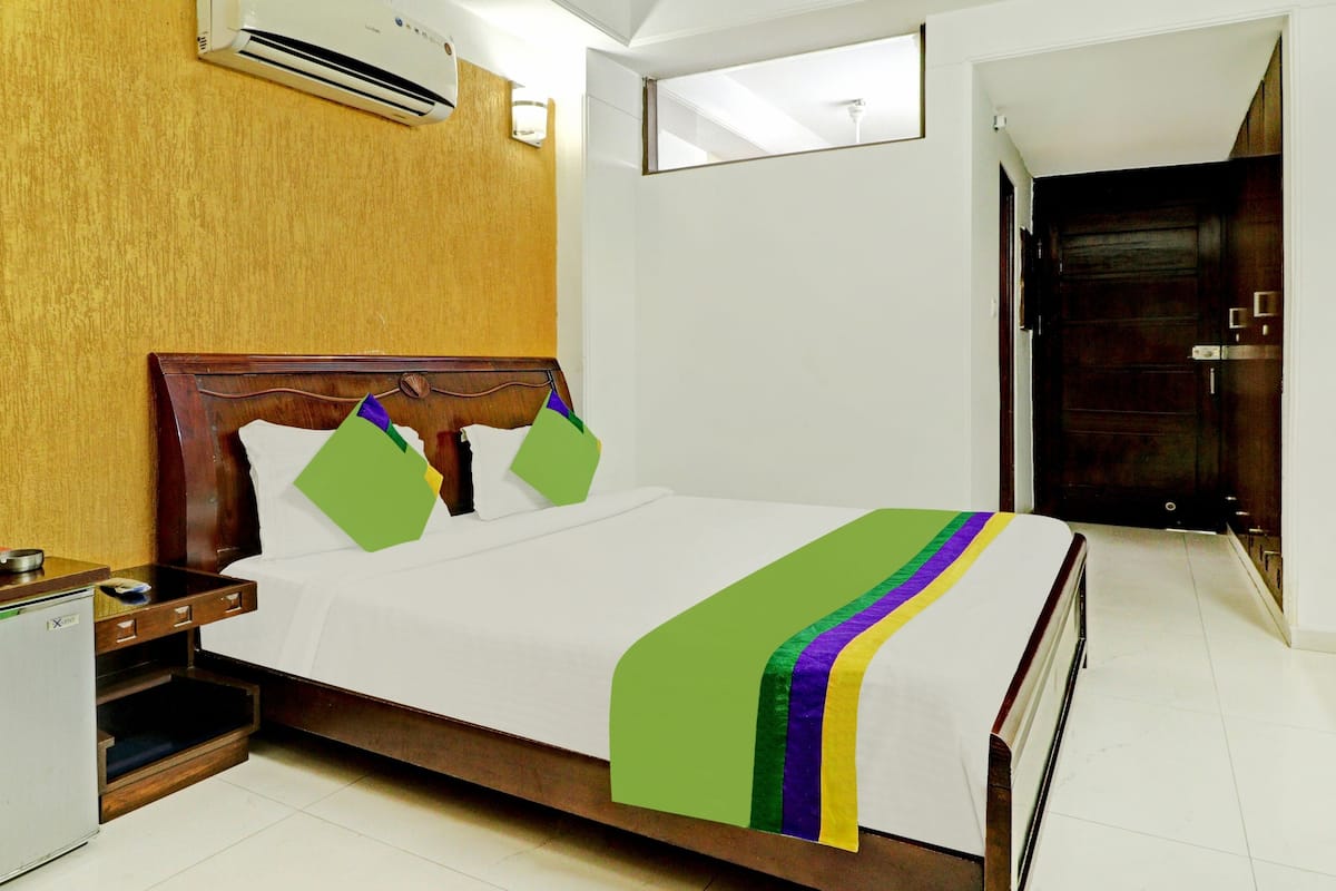 Quarto de hotel com cabeceira de madeira, cama com lençol branco e manta colorida, mesa de cabeceira pequena com abajur e ar-condicionado fixado na parede.