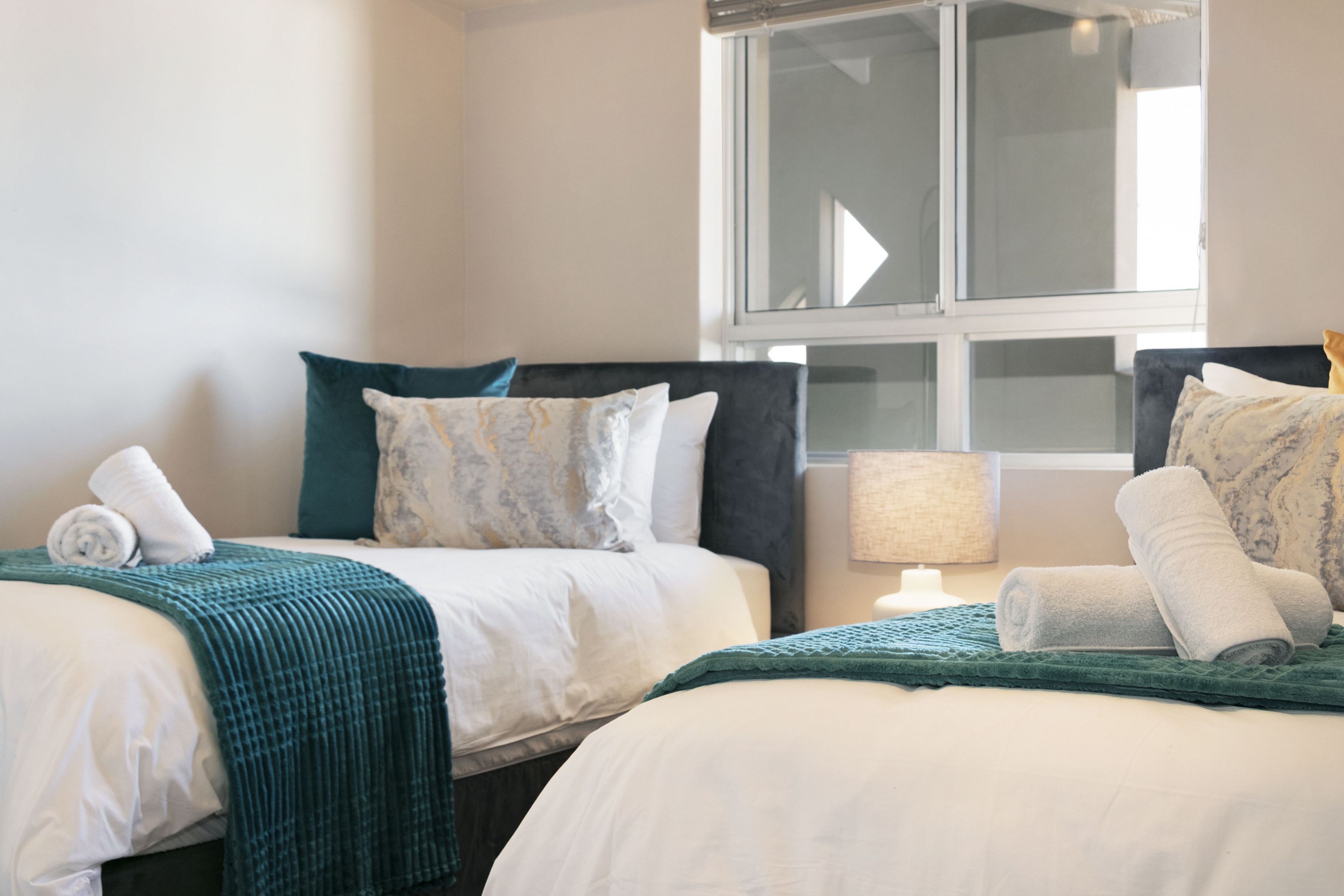 A hotel room with two beds, each with a green blanket and white linens. There is a window with a view of the outside.
