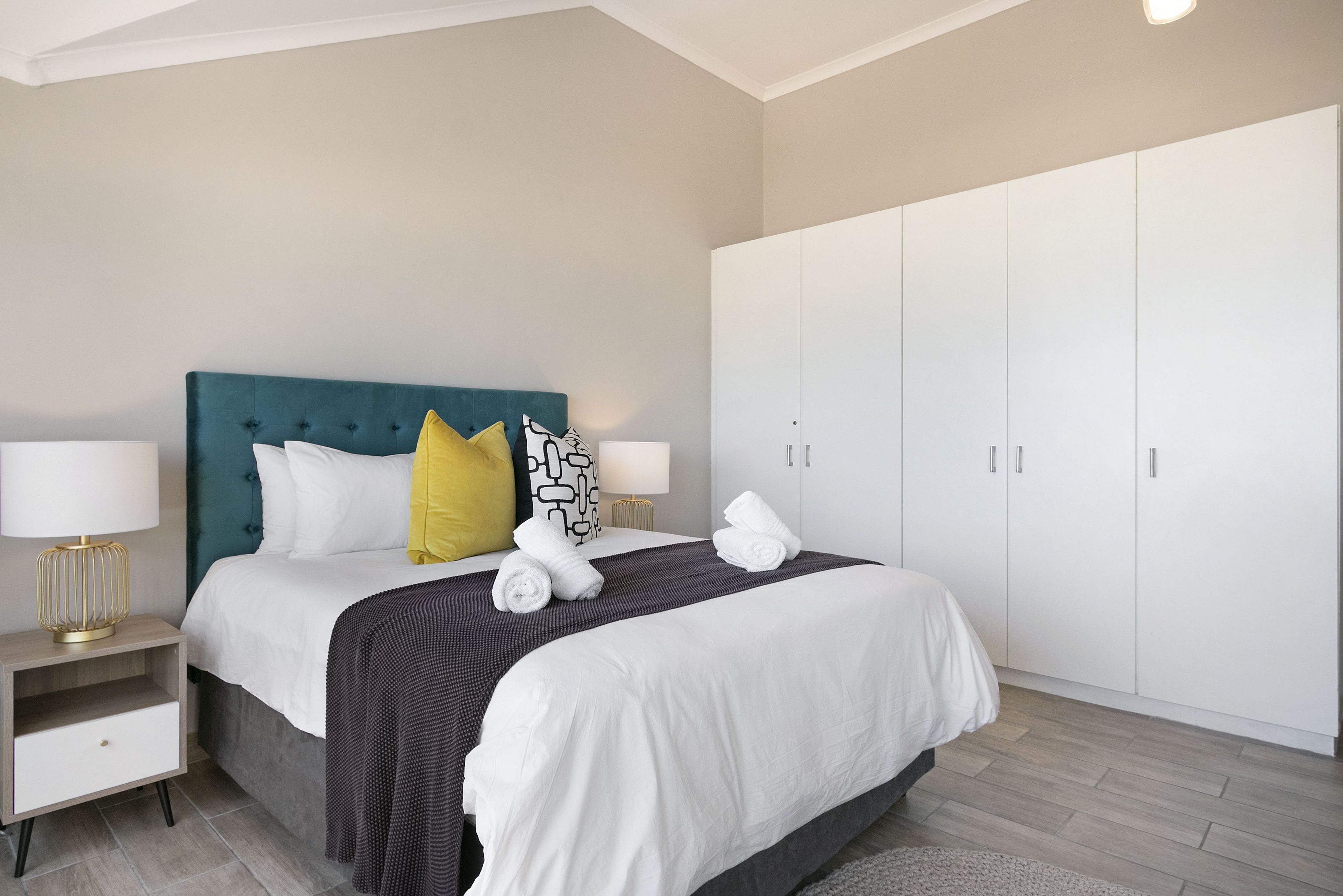 A neatly made bed with white linens, a teal headboard, and decorative pillows. There are two bedside tables with lamps, and a large wardrobe with sliding doors.