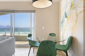 A modern room with a glass table and chairs, a sofa, a large abstract painting, and a view of the ocean and mountains. - Sea Spray B404 by HostAgents (Cape Town)