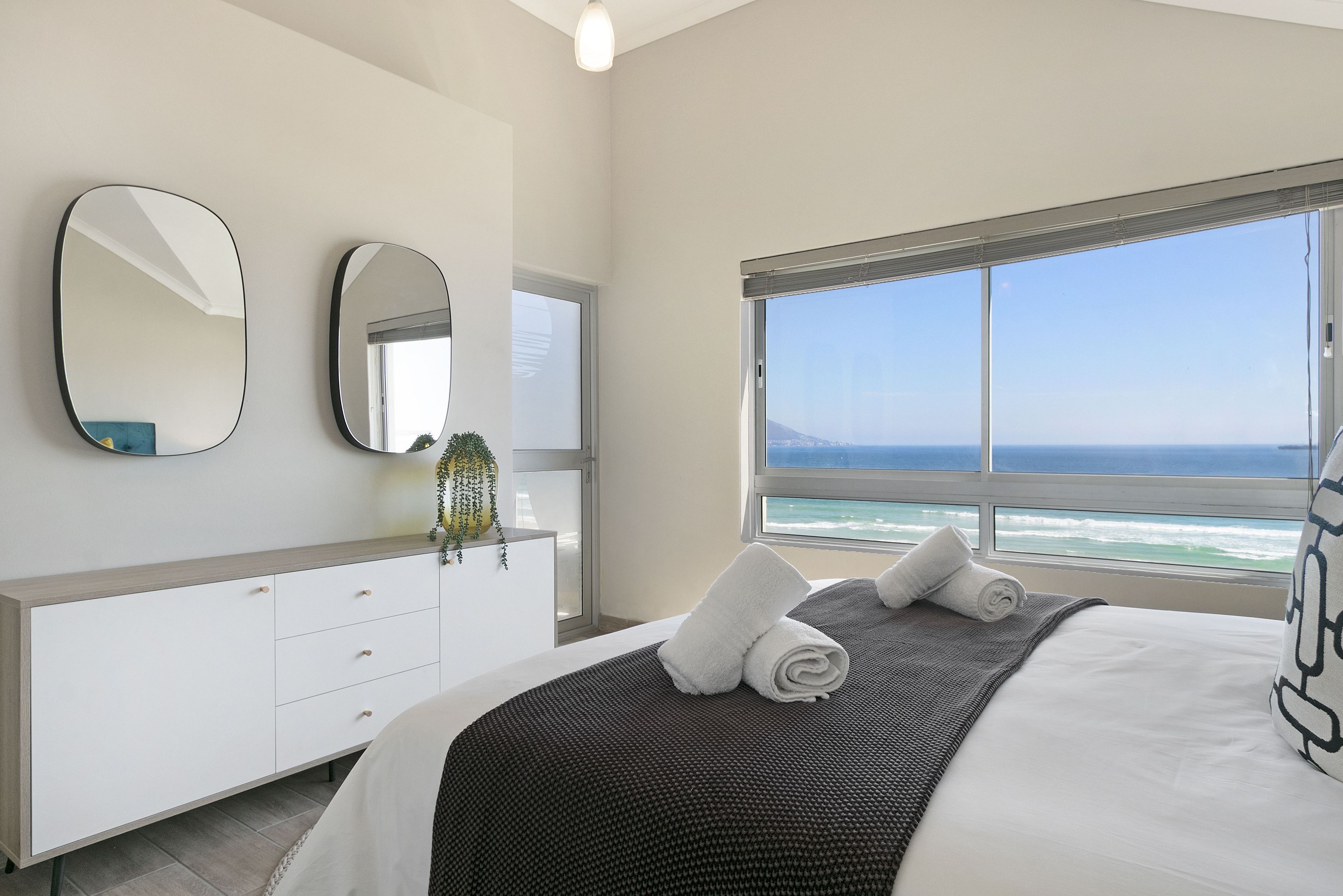 A modern bedroom with a large bed, a dresser, two mirrors, and a view of the ocean.