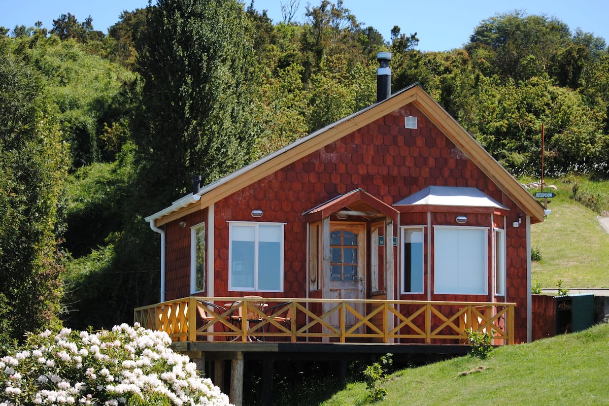 A red wooden cabin with a balcony, surrounded by lush greenery.