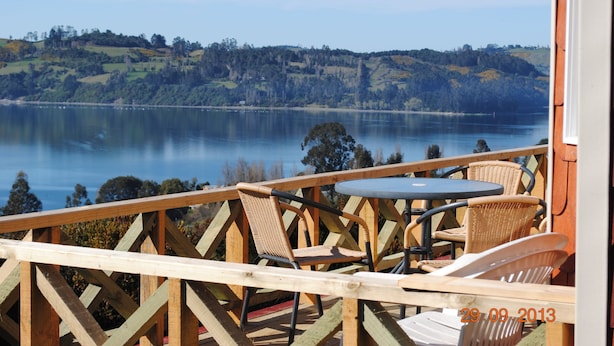 A wooden deck with wicker chairs and a table overlooking a lake with hills in the background. - Cabañas Trayen Chiloé (Castro)