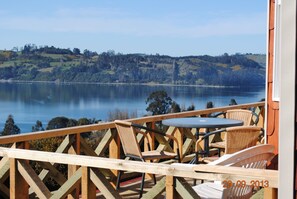 A wooden deck with wicker chairs and a table overlooking a lake with hills in the background. - Cabañas Trayen Chiloé (Castro)