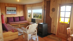 A cozy room with a bed, a chair, a desk, a fireplace, and large windows offering a view. - Cabañas Trayen Chiloé (Castro)