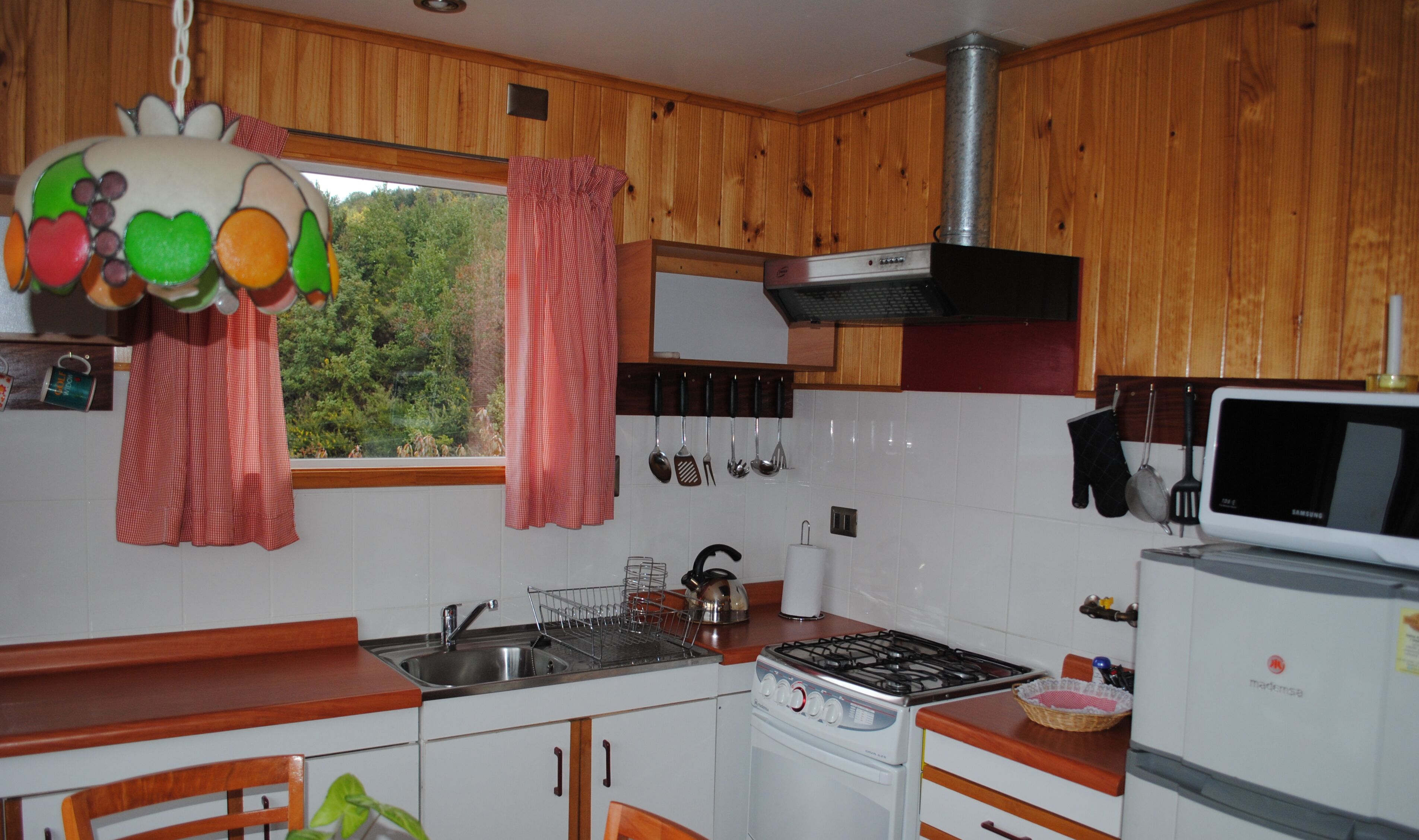Una cocina con armarios de madera, una estufa a gas, un microondas y una ventana con cortinas.