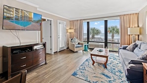 A hotel room with a television, a sofa, a coffee table, a chair, a window with a view of the beach, and a large window with curtains.