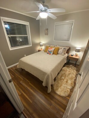 Quarto com cama, ventoinha no teto, duas mesas de cabeceira com luminárias, tapete fofinho e janela com persianas.