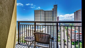 A balcony with a wicker chair and a table, overlooking a beach and cityscape with tall buildings.