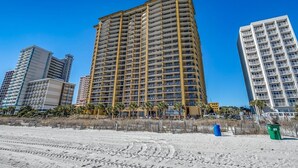A high-rise hotel with multiple floors, a sandy beach in the foreground, and other buildings in the background.