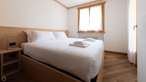 A neatly made bed with white linens and folded towels, a wooden nightstand with a lamp, and a window with curtains. - Castellazzi 12 Aquila (Bormio)