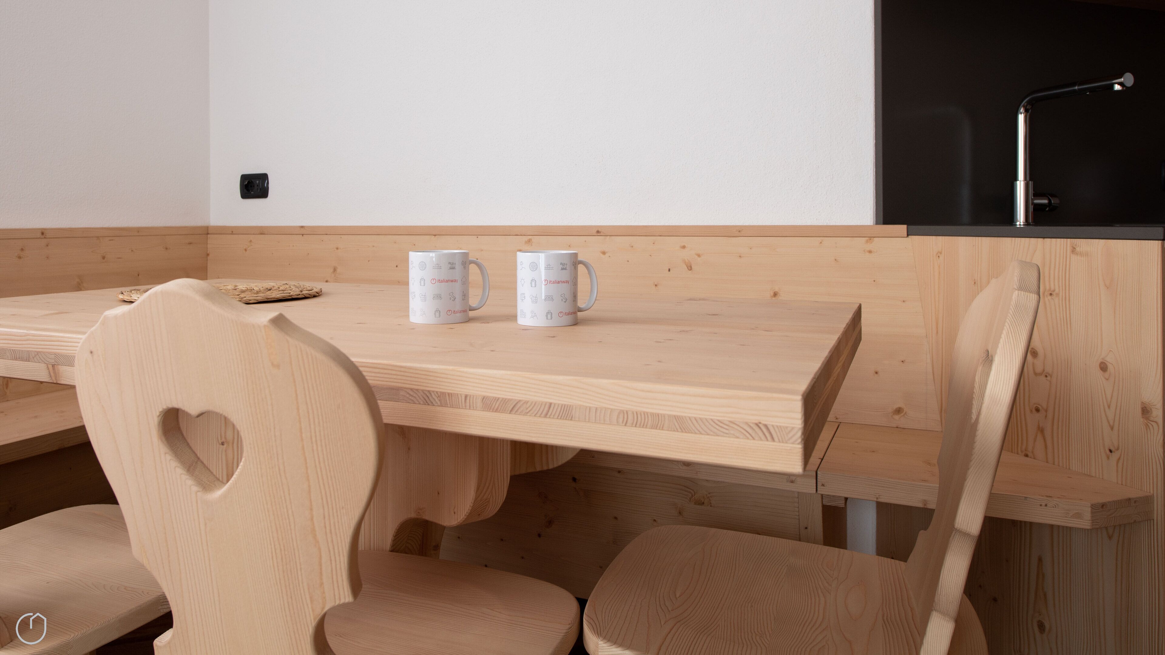 A wooden dining table with two white mugs, a heart-shaped backrest chair, and a black faucet in the background.
