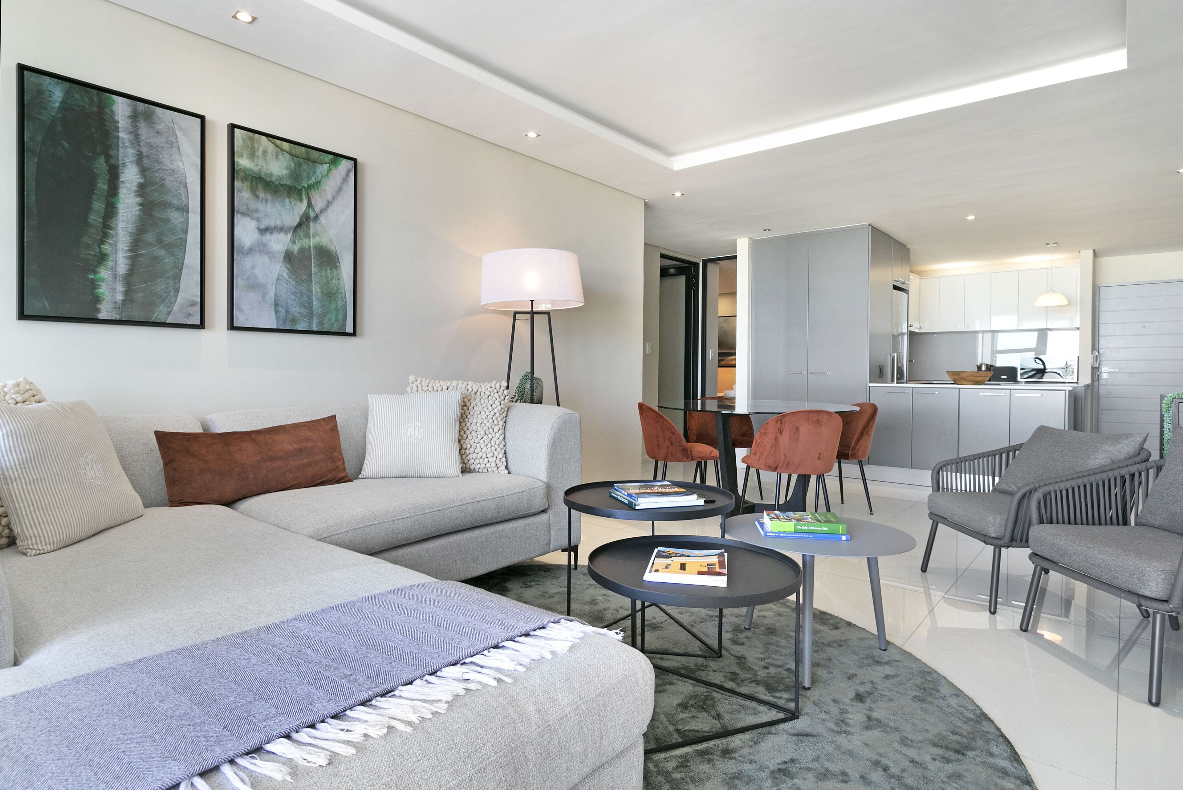 A modern living room with a sofa, coffee table, and chairs, a kitchen in the background, and artwork on the wall.